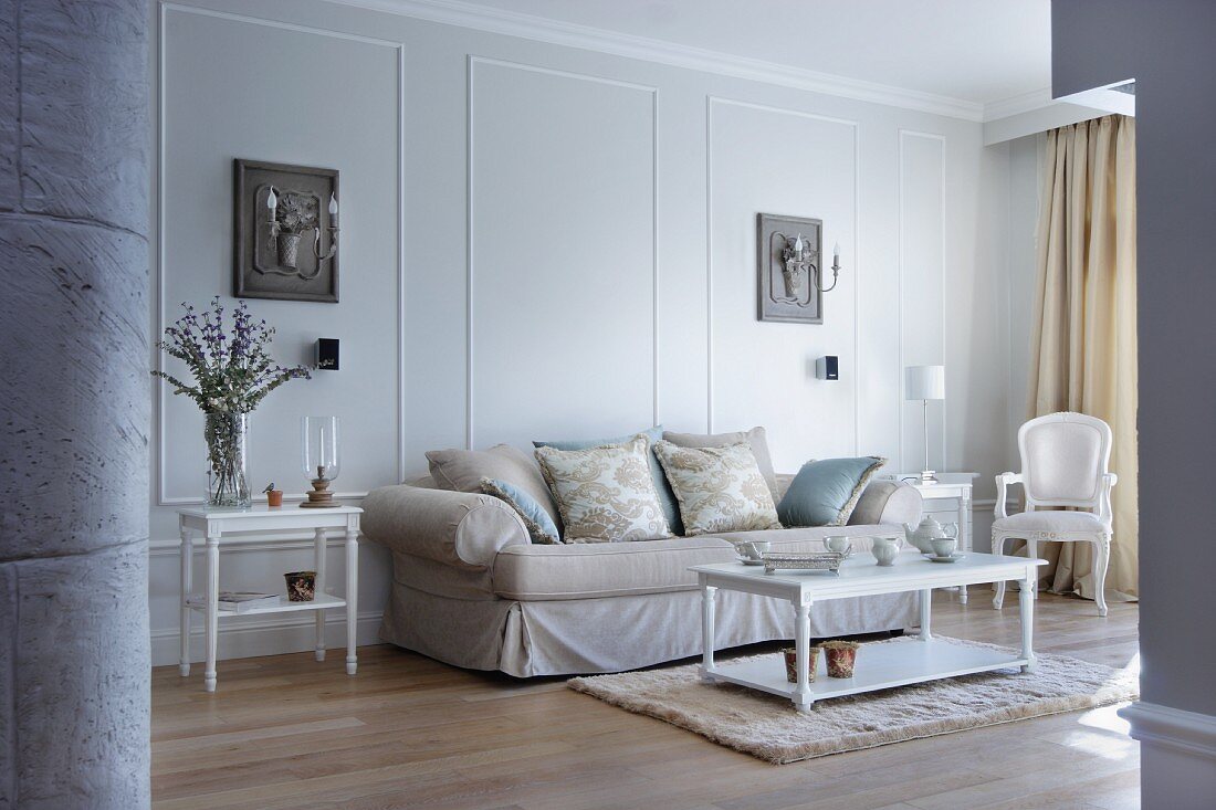 Elegantes Sofa und weiße antikisierende Möbel vor lichtgrauer Wand mit Stuckrahmen und Leuchter-Kassetten