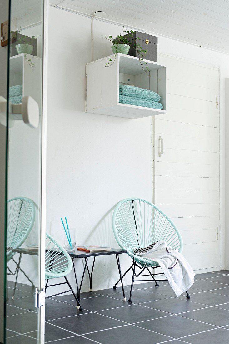 Retro Sessel mit türkiser Seilverspannung auf grauem Fliesenboden, vor Wand mit aufgehängtem Regalmodul