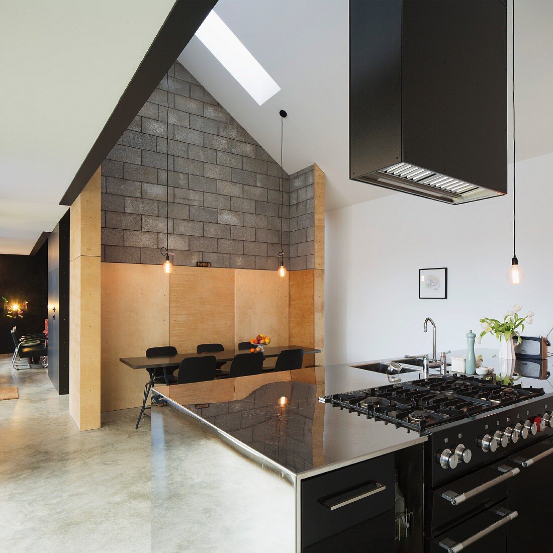 Stainless steel kitchen counter under extractor hood, dining area in wood-panelled niche and concrete block wall in modern, open-plan interior