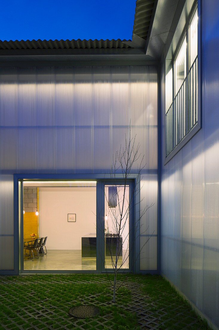 Twilight atmosphere in courtyard of modern house; view through illuminated, floor-to-ceiling terrace door into open-plan interior