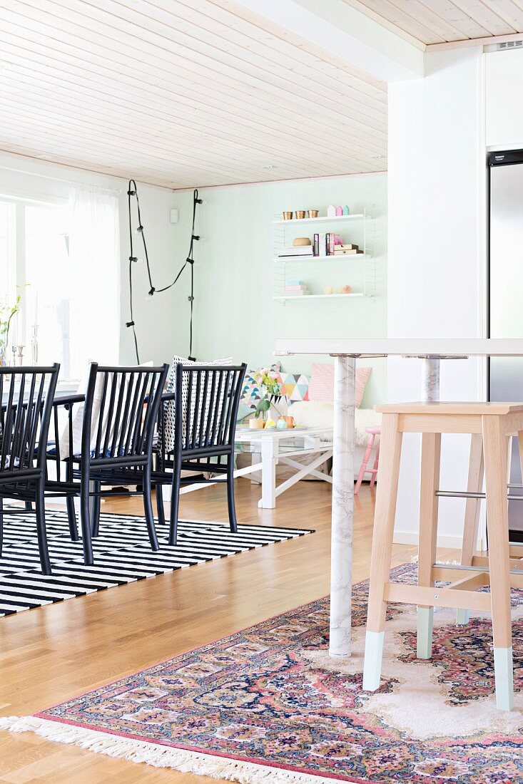 Offener Wohnraum mit Holzdecke, vorne teilweise sichtbare Theke und Holz Barhocker, im Hintergrund schwarze Stühle an Esstisch