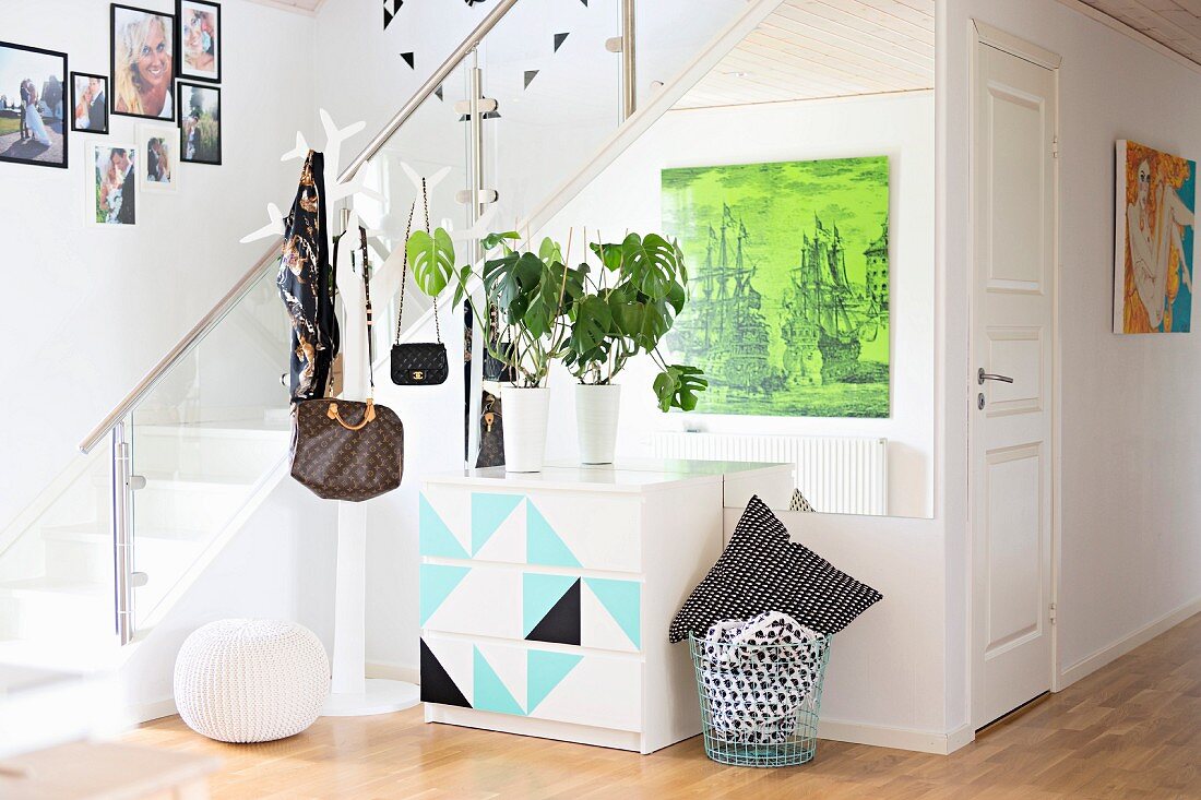 Grünpflanzen auf grafisch gestalteter Kommode und Garderobenständer mit aufgehängten Taschen, neben Treppe in modernem Gangbereich