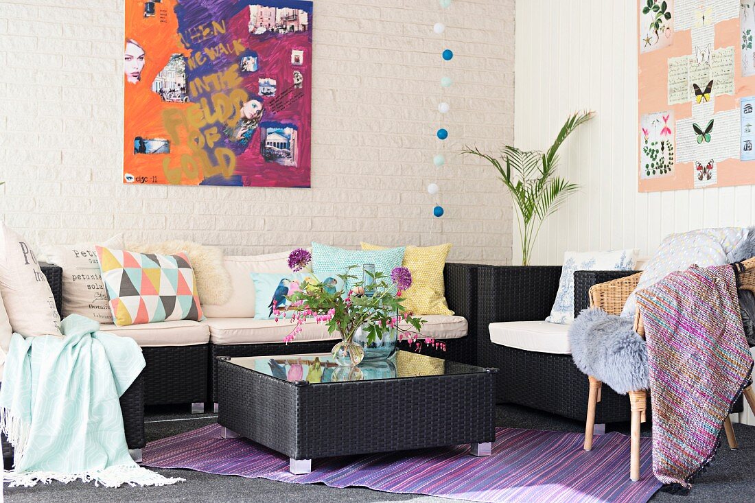 Sofa mit verschiedenen Kissen und Coffee Table, aus dunklem Rattangeflecht, in Zimmerecke, vor geweisselter Wand mit modernem Bild