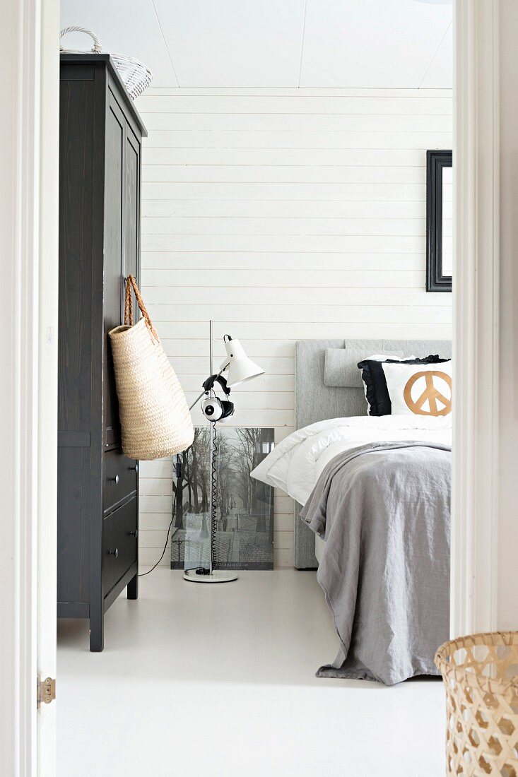 Blick durch offene Tür ins Schlafzimmer, seitlich schwarz lackierter Kleiderschrank und Stehleuchte neben Bett mit grauer Tagesdecke