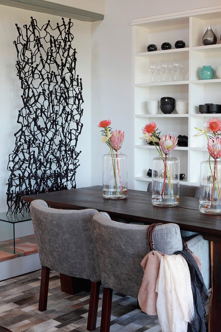 Gepolsterte Stühle mit hellgrauem Bezug an dunklem Holztisch, darauf Vasen mit Protea Blume in Reihe, in Zimmerecke