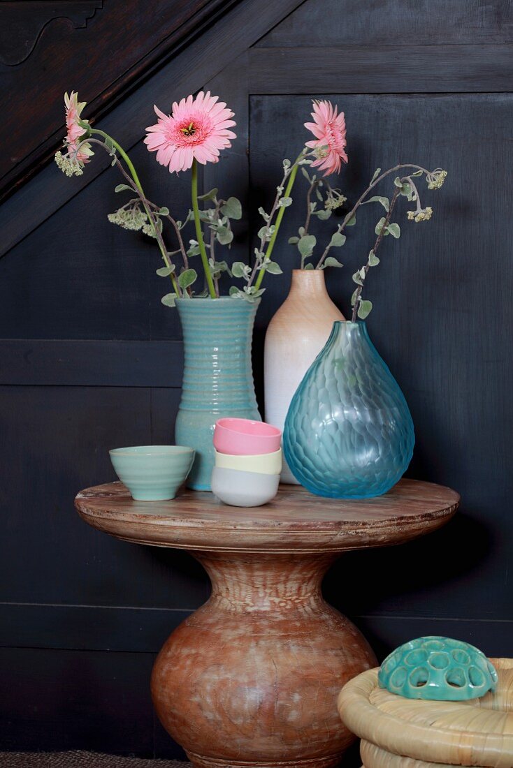 Verschiedene Vasen mit rosa Gerberas und pastellfarbene Schälchen auf rundem Steintisch