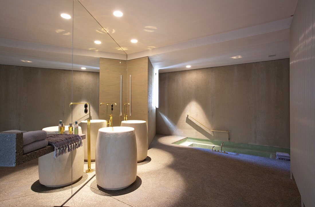 Cylindrical pedestal sink with brass floor-mounted taps against mirrored wall in luxurious bathroom with sunken bathtub