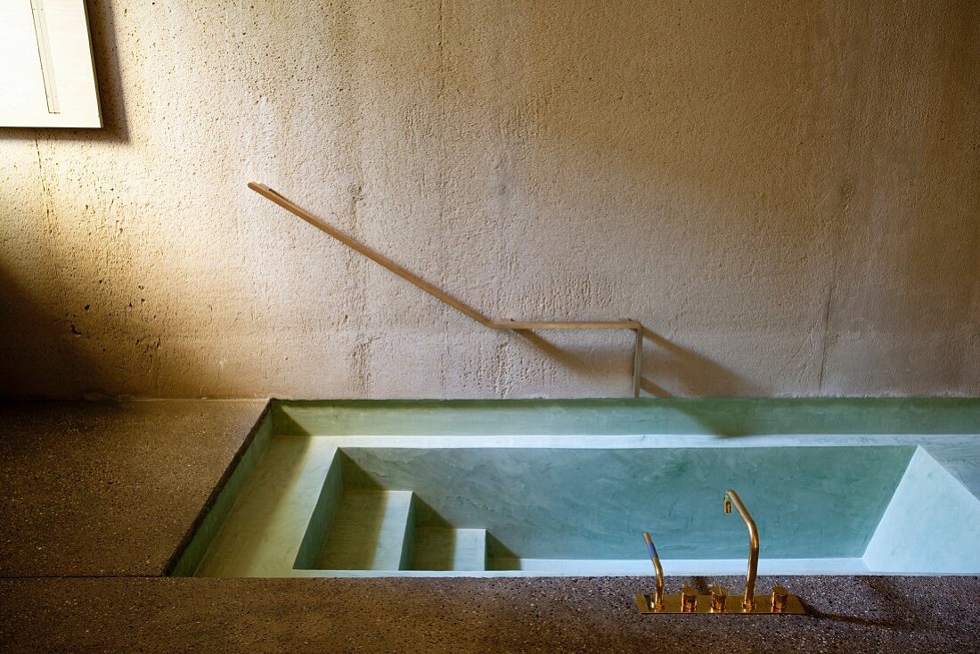 Sunken concrete bathtub with brass taps and minimalist handrail on concrete wall