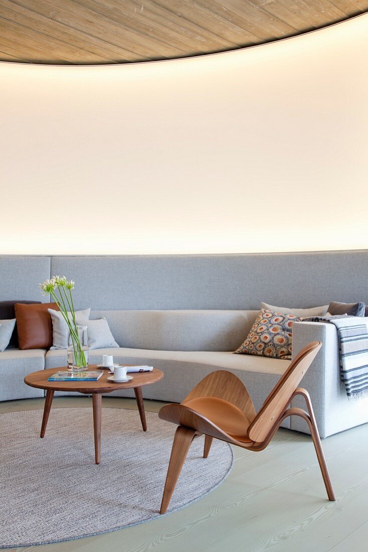 Klassikerstuhl und Coffeetable aus Holz auf rundem Teppich, vor hellgrauem, massgefertigtem Sofa an gebogener Wand