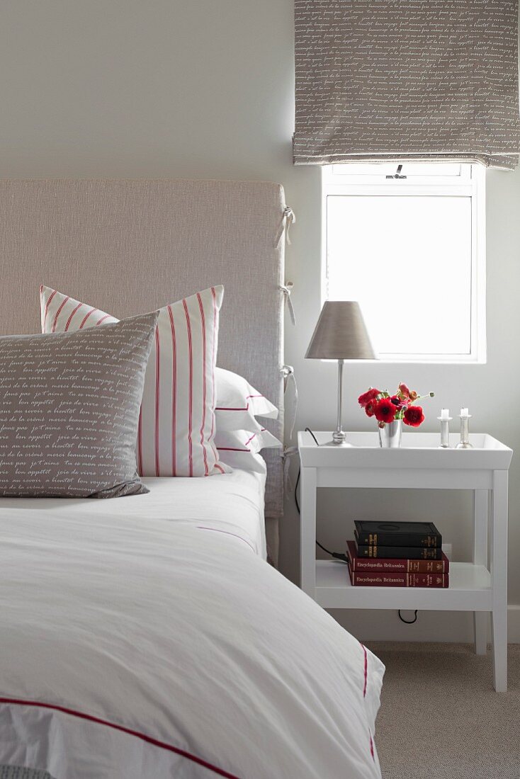 Bedside table below window with Roman blind and scatter cushions on bed in white bedroom
