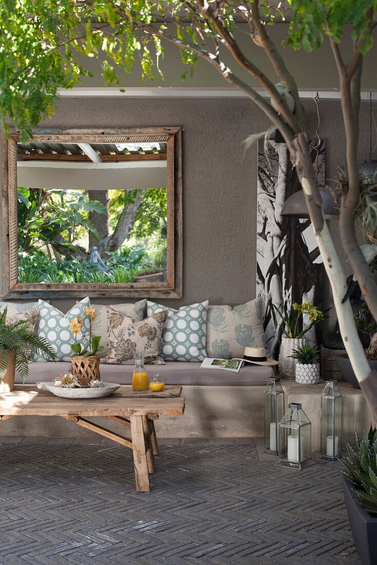 Sitzecke mit Kissen vor grauer Wand mit Spiegel auf Terrasse im sommerlichen Garten, Trompe l'oeil