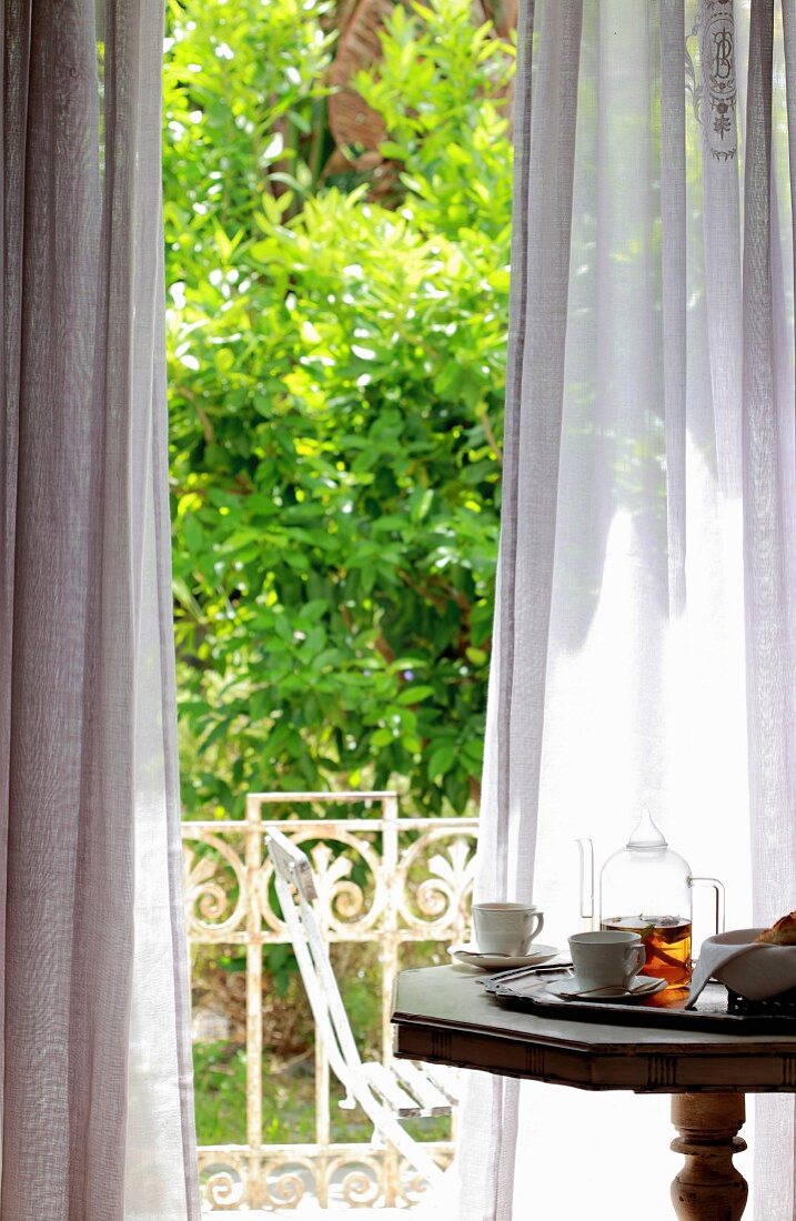 View across breakfast table though wafting curtains into green garden