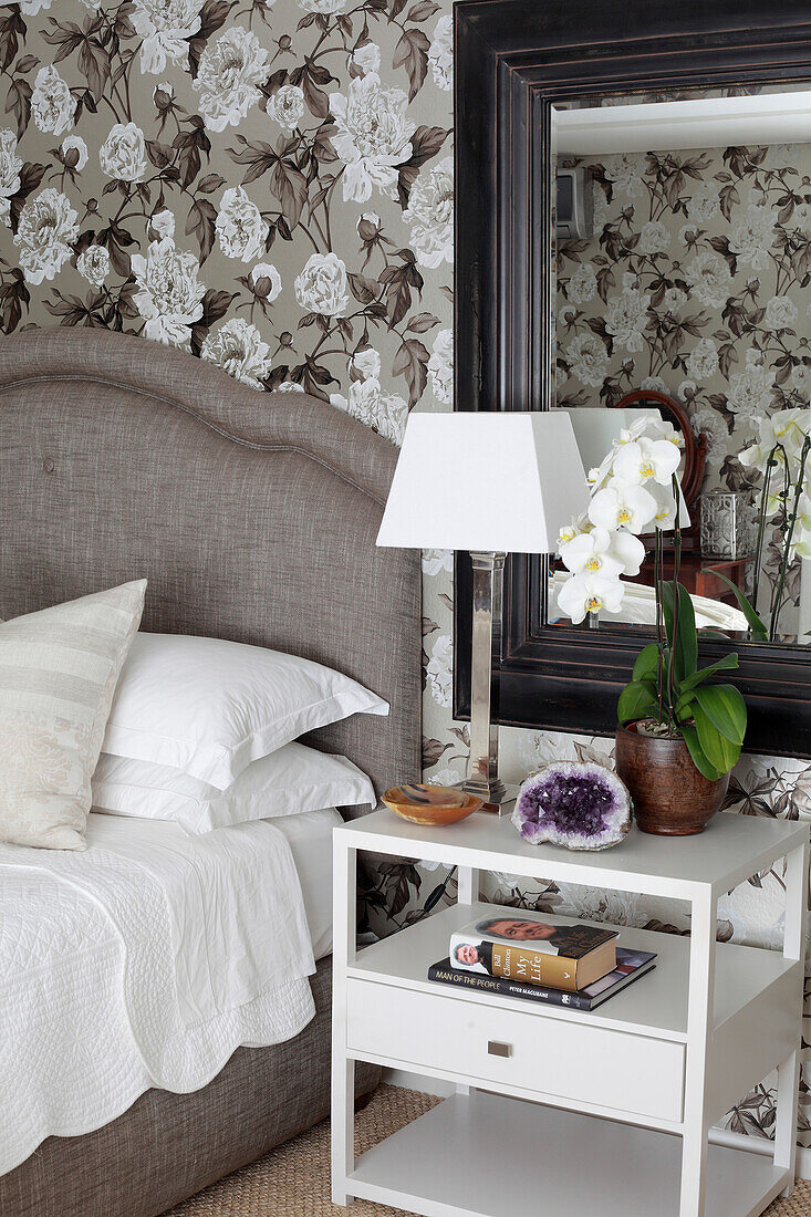 Table lamp and orchid on white bedside table with drawer below framed mirror and next to bed in elegant bedroom