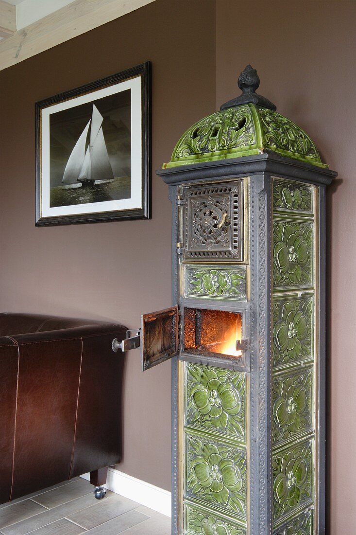 Antique, Art Nouveau, wood-burning stove with green tiles against brown-painted wall; leather armchair below picture of sailing boat to one side
