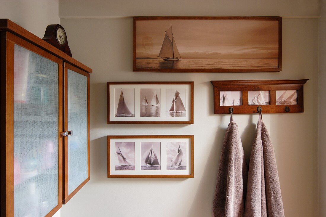 Gerahmte Fotos mit Segelboot Motiven und Handtücher auf Hakenleiste an Wand, seitlich Hängeschränkchen aus Holz mit opaken Glastüren