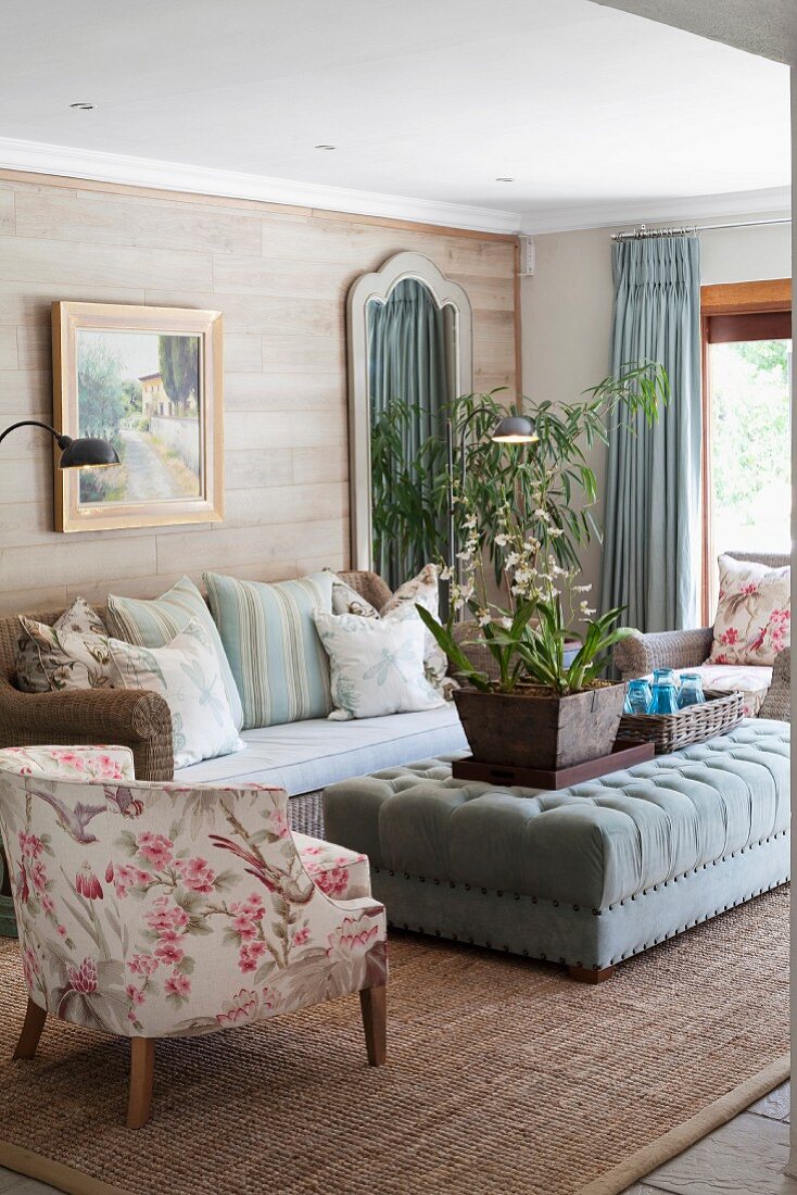 Pastel living room with wicker sofa against wooden wall, velvet ottoman, floral armchair and jute rug