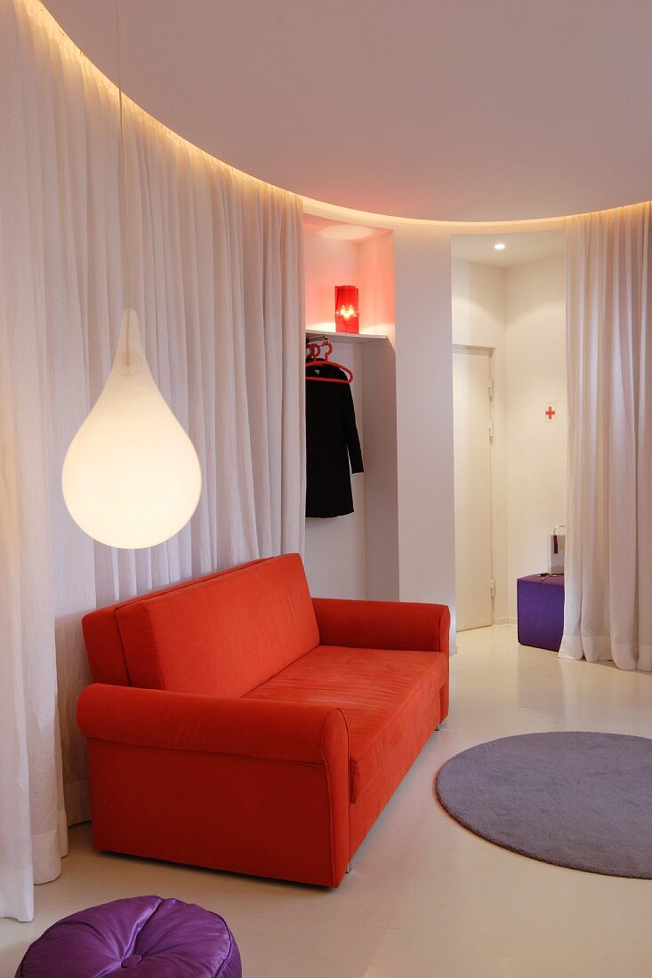 Tropfenförmige Hängeleuchte und orangerotes Sofa vor weißem Vorhang als Raumabtrennung in minimalistischem, kreisförmigem Loungebereich