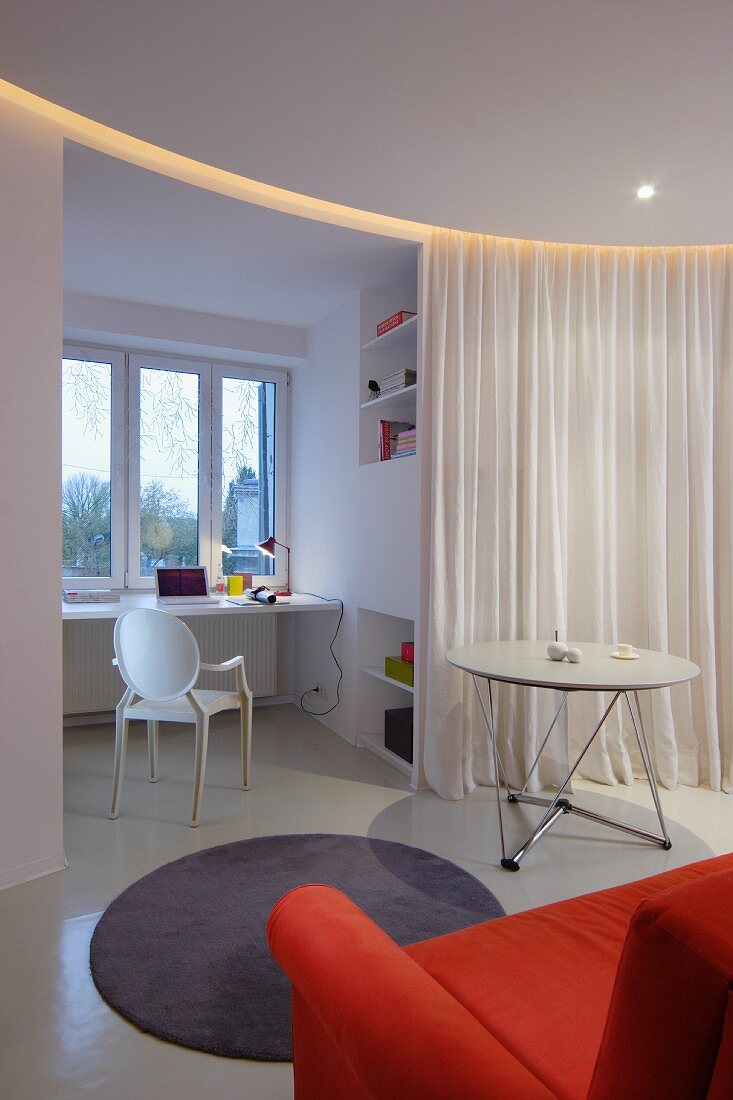 Weisser Tisch und runder grauer Teppich in abgetrenntem Wohnbereich, offener Vorhang mit Blick auf Arbeitsecke mit weißem Ghoststuhl vor Fenster