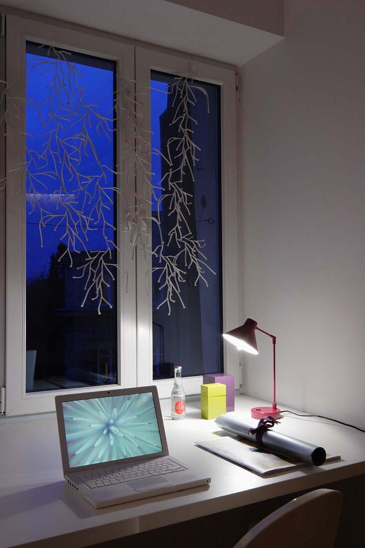 Laptop neben Papierrolle und Schreibtischleuchte auf Tisch am dekorierten Fenster mit Dämmerungsstimmung
