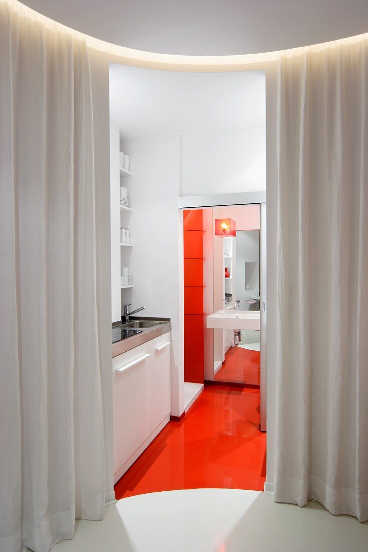 Blick durch geöffneten Vorhang in moderne Miniküche mit orangerotem Boden, hinter offener Schiebetür Blick ins Bad