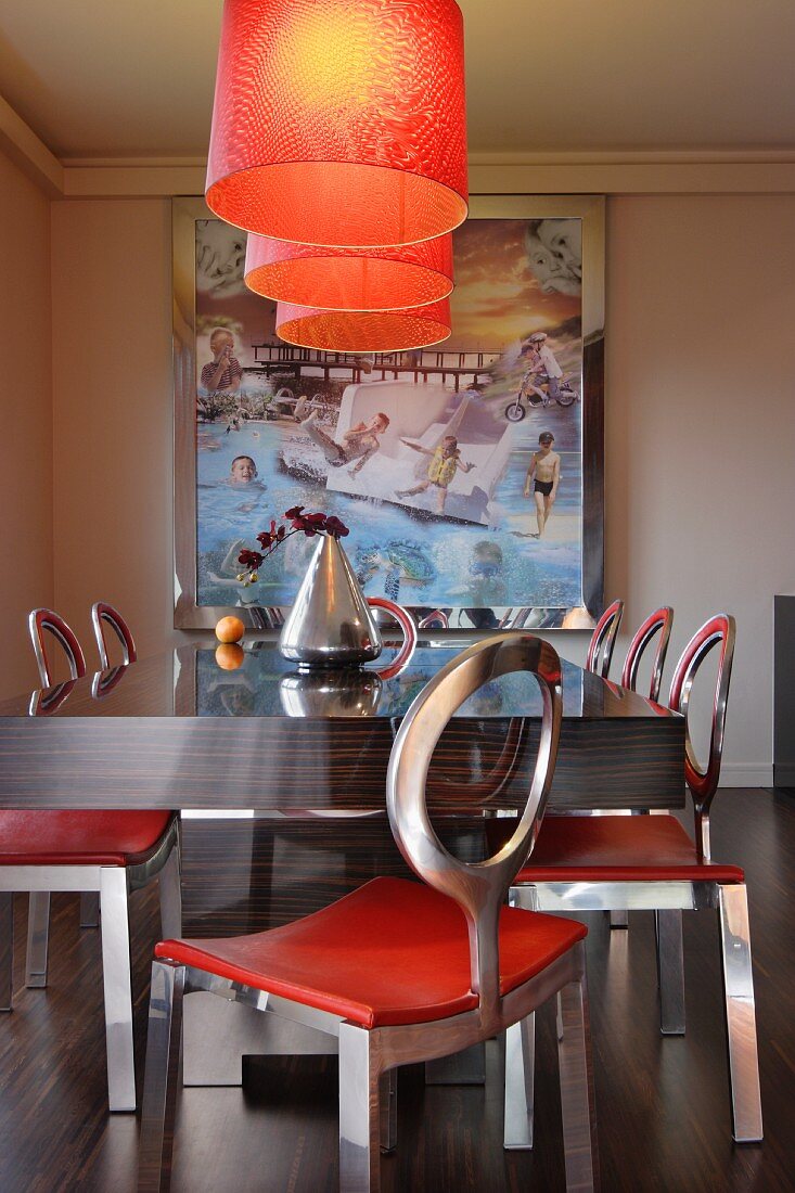 Orange lamps above dark, exotic wood table and metal chairs with circular backrests; surreal artwork in background