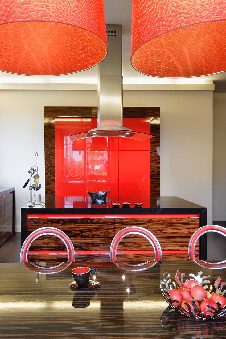 Orange lamps above dark exotic wood table and metal chairs with circular backrests in open-plan exotic wood kitchen with red fitted elements