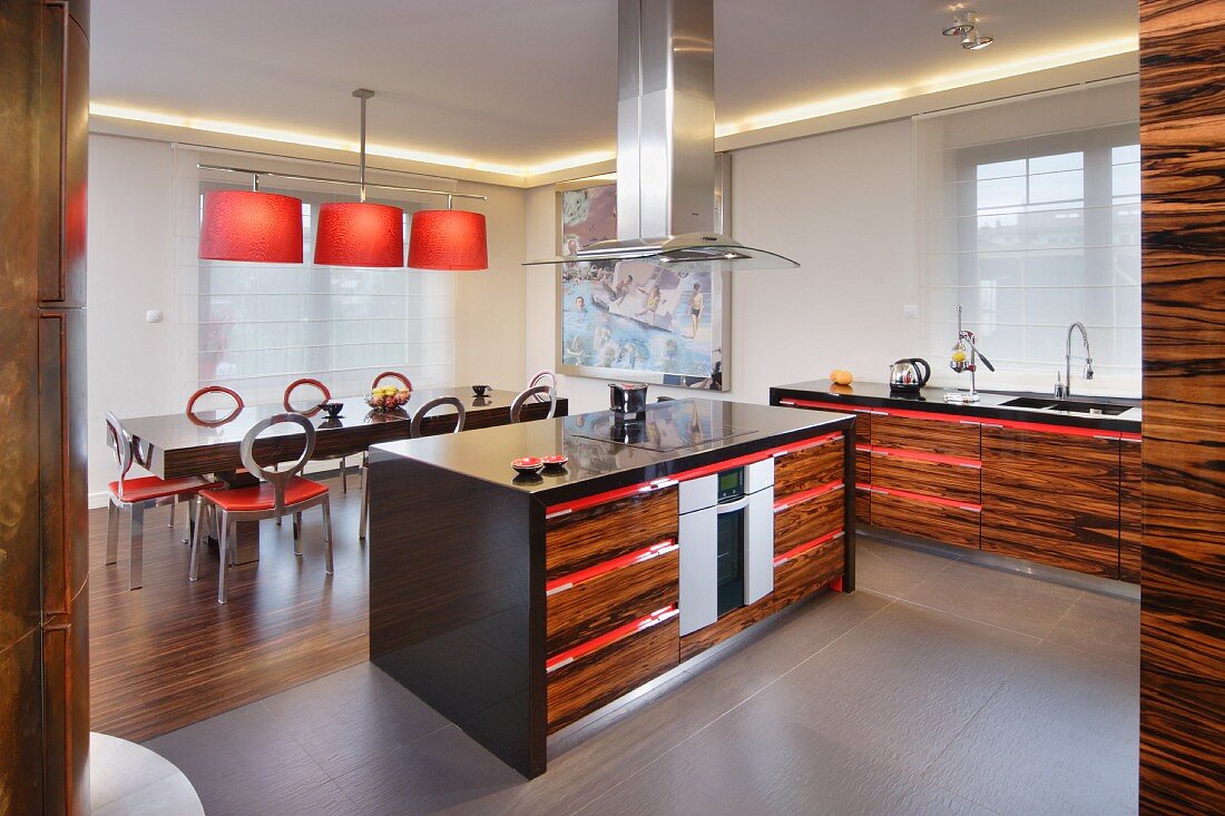 Herdinsel in offener Küche mit Edelholzfronten und roten Akzenten in den Griffleisten; farbharmonisch abgestimmter Essplatz im Hintergrund