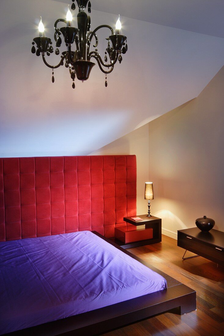 Niedriges, dunkles Holzbett mit hellviolettem Laken, rote Polsterwand und Kronleuchter in kleinem Dachzimmer
