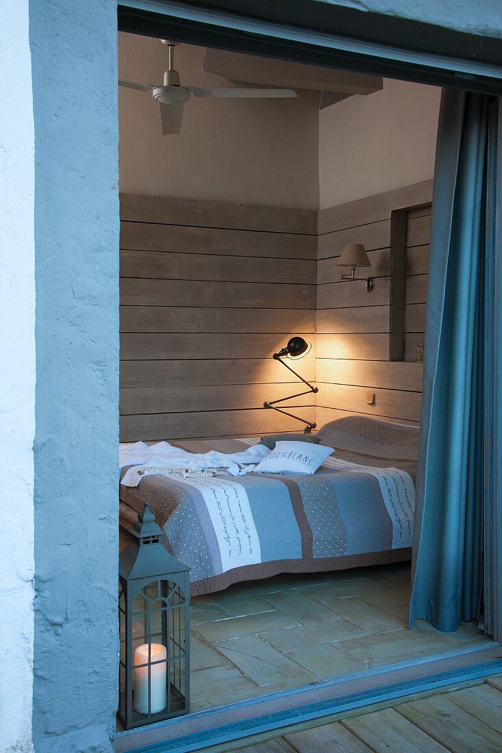 View through open terrace door into wood-clad bedroom with retro table lamp in corner