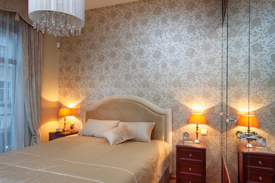 Französisches Bett mit Polster Kopfteil und Nachtschränkchen mit Tischleuchte, vor Wand mit heller Seidentapete in traditionellem Schlafzimmer