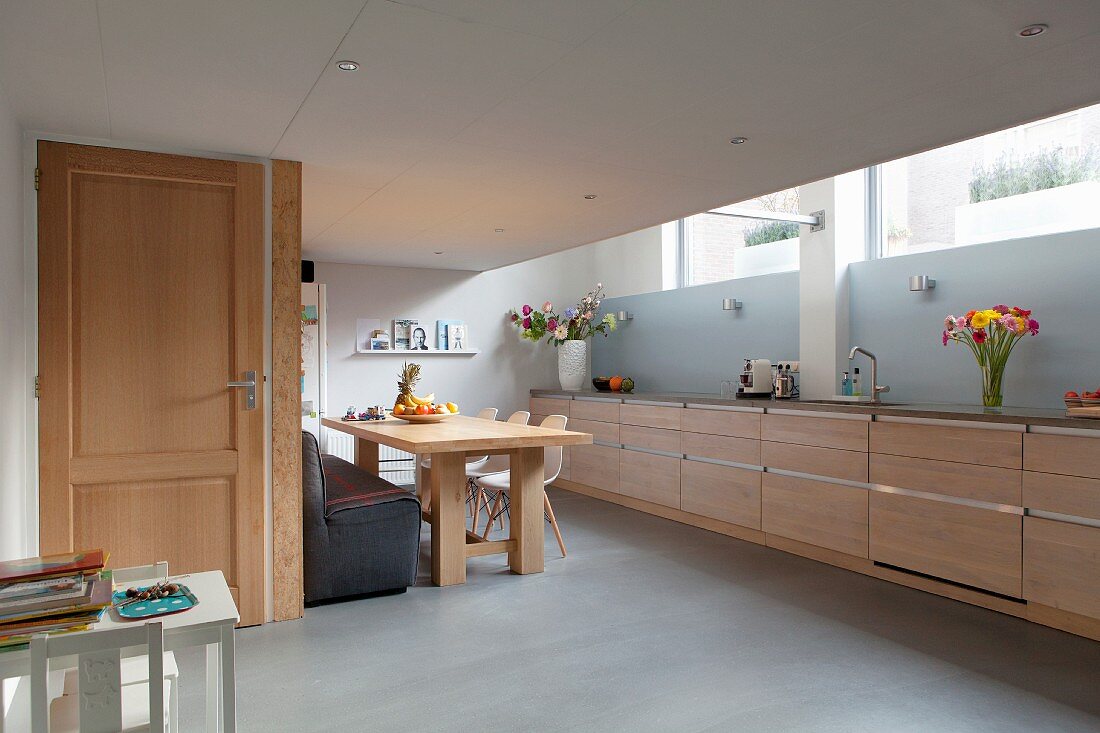 Elongated kitchen counter with pale wooden fronts and pale blue splashback in kitchen dining room with white children's table and kitchen door