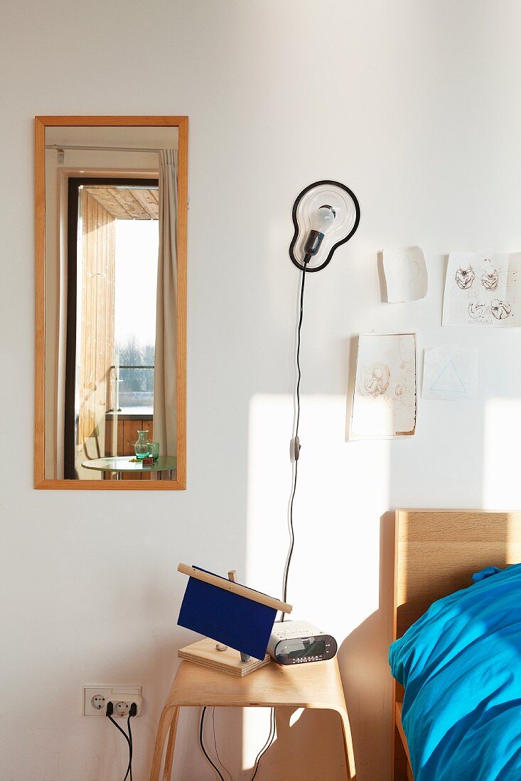 Moderner Nachttisch aus Holz und Wandleuchte zwischen aufgehängtem Spiegel und Bett
