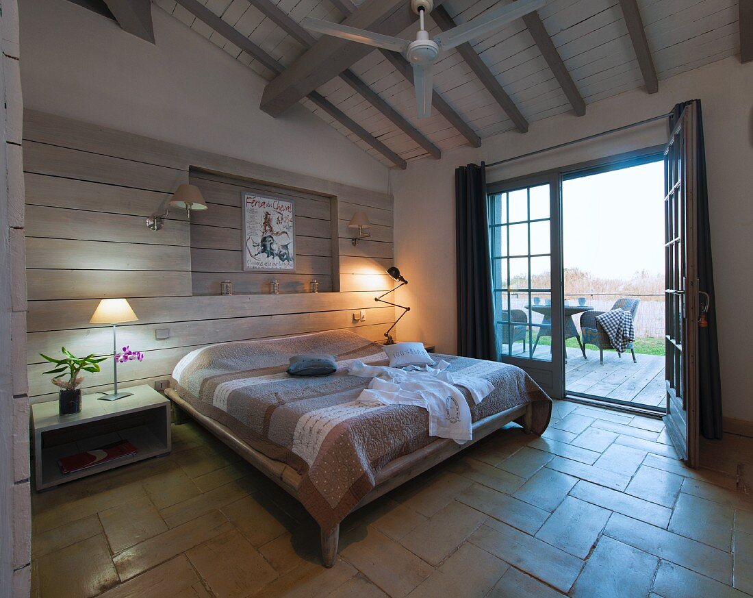 Doppelbett vor holzverschalter Wand, zwischen Nachttischen mit Tischleuchten in mediterranem Schlafzimmer, offene Terrassentür mit Ausblick