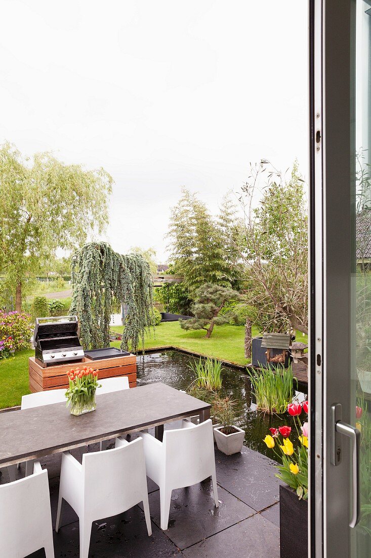 Blick von Terrasse mit modernem Edelstahl-Chassis Sitzplatz und Outdoor-Küche auf Teich