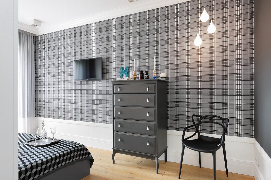 Dark grey chest of drawers and black chair against grey and black tartan wallpaper in bedroom