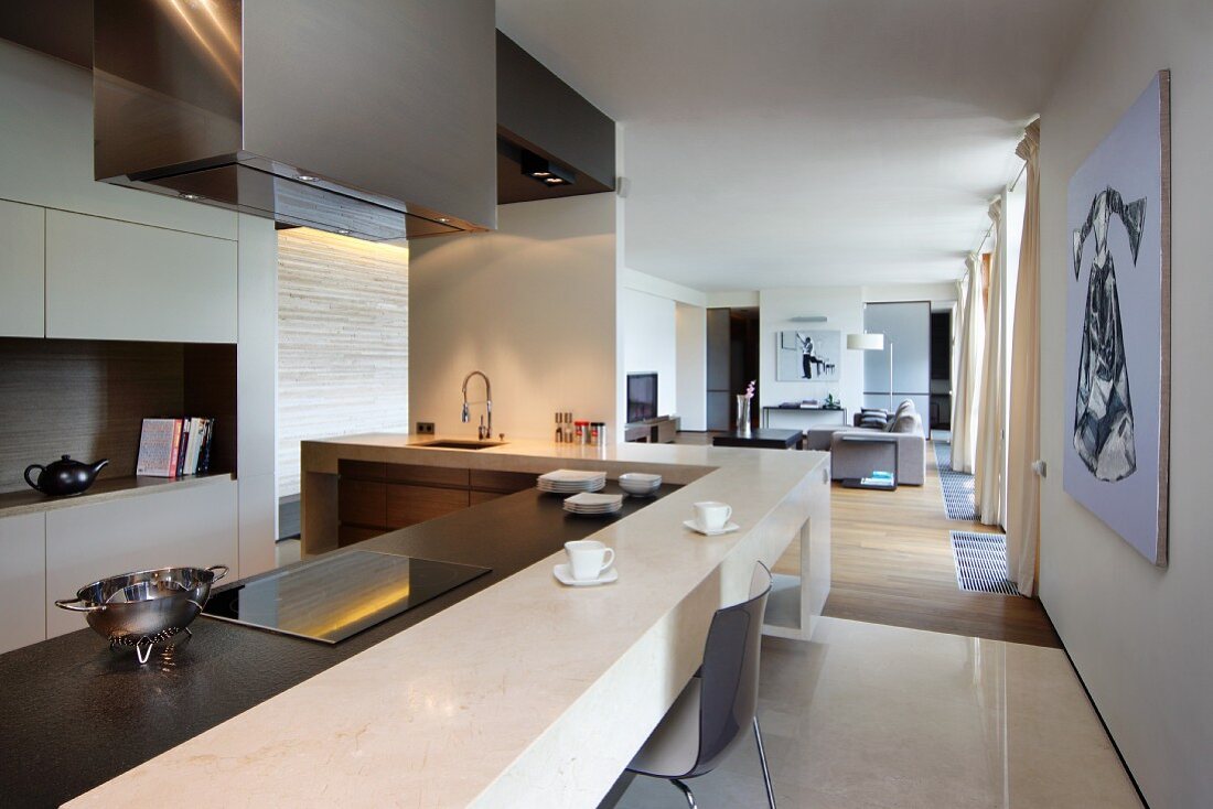 Designer Küche mit Frühstückstresen an L-förmiger Arbeitsfläche; im Hintergrund ein langgestreckter Wohnraum in Naturtönen