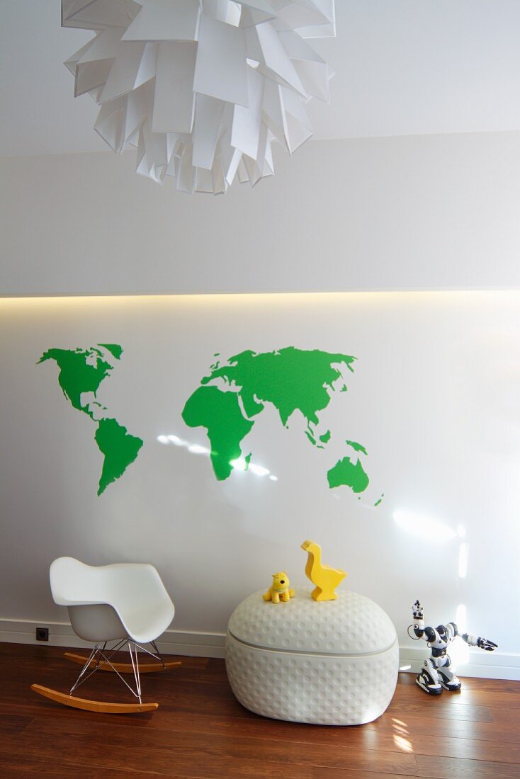 Kinder-Schaukelstuhlklassiker und Spielzeug auf Designertruhe vor einer Wand mit indirekter Beleuchtung und Aufdruck einer grünen Weltkarte