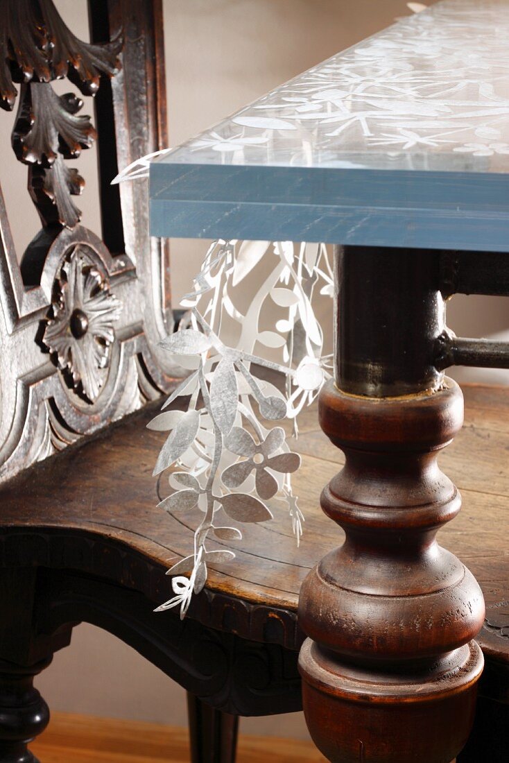 Aus Stoff ausgeschnittene florale Motive zwischen zwei Glasplatten auf Stahlkonstruktion mit gedrechselten Holzbeinen als kreatives Tischmodell mit antikem Holzstuhl