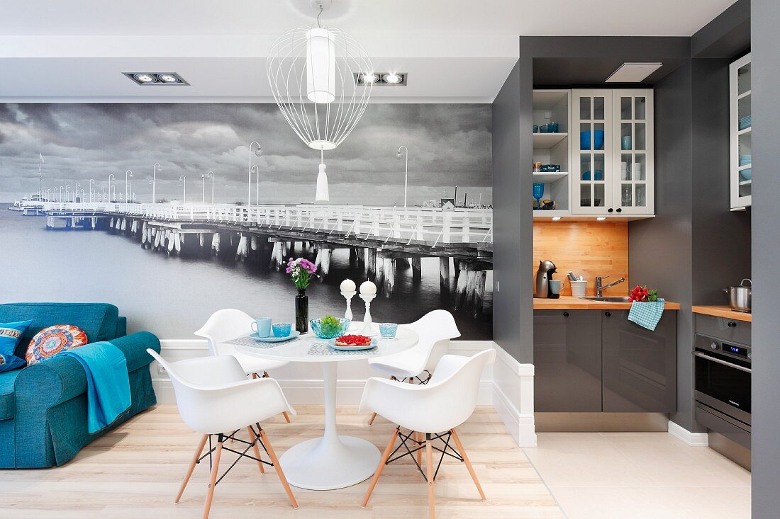 Tulpentisch mit Plastic Armchairs und ein blaues Sofa vor schwarz-weisser Fototapete; seitllich die offene, grau dominierte Designerküche