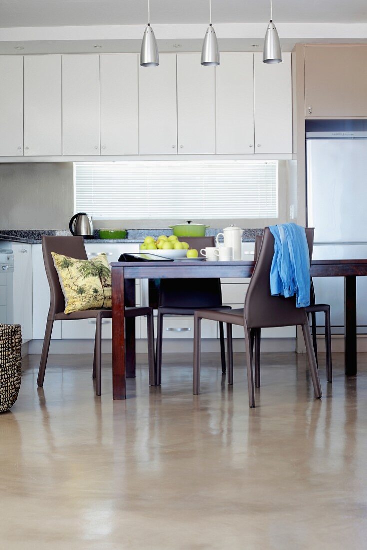 Dunkler Holzesstisch und graue Kunststoff Stühle auf poliertem Estrichboden vor Küchenzeile mit weissen Einbauschränken in modernem Ambiente