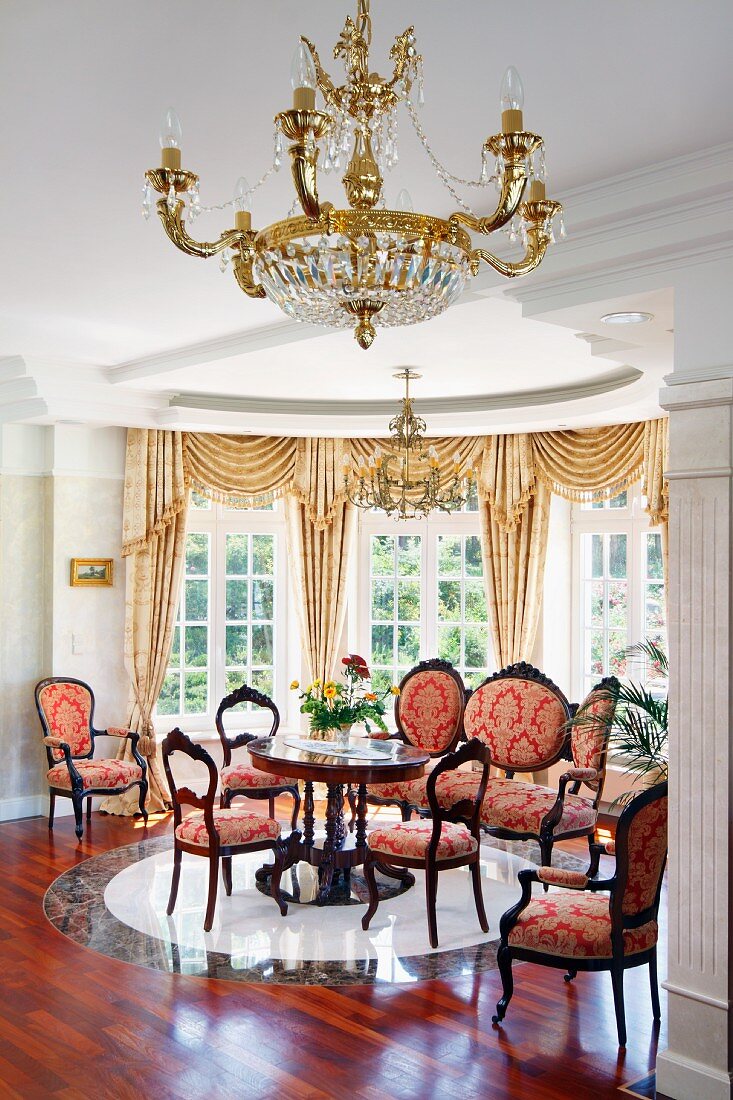 Goldener Kronleuchter vor Salonbereich mit Stilmöbeln, Stühle mit rotgoldenem Ornamentmuster auf Stoffbezug, in halbkreisförmigem Erker in herrschaftlichem Ambiente