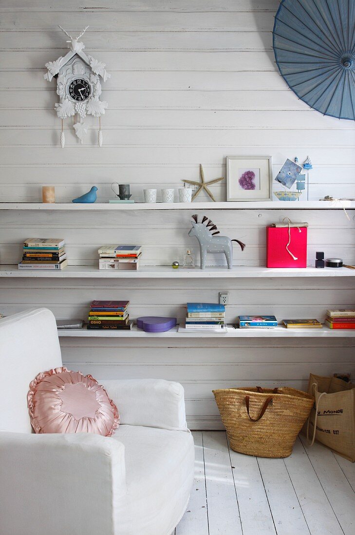 White armchair in front of white wooden wall with books & ornaments on shelves below old cuckoo clock