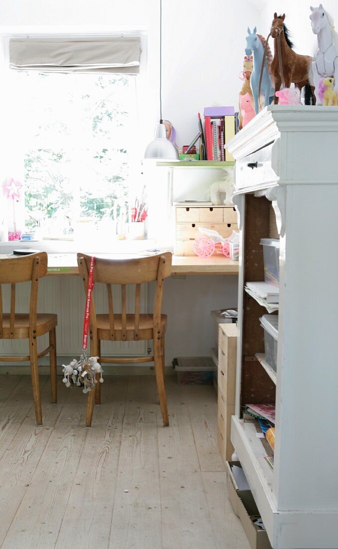 Schlichte Holzstühle an Schreibtisch vor Fenster, seitlich Pferdefiguren auf offenem Schrank, weiss lackiert, im Kinderzimmer