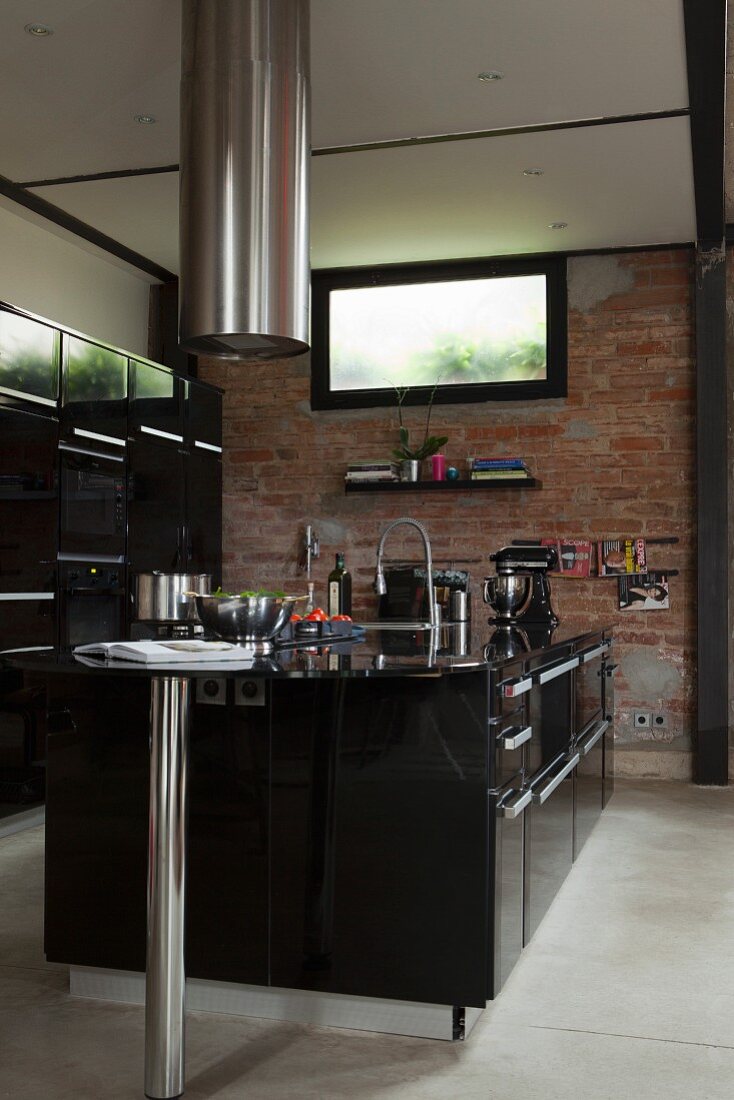 Black free-standing designer kitchen counter below cylindrical stainless steel extractor hood in front of brick wall