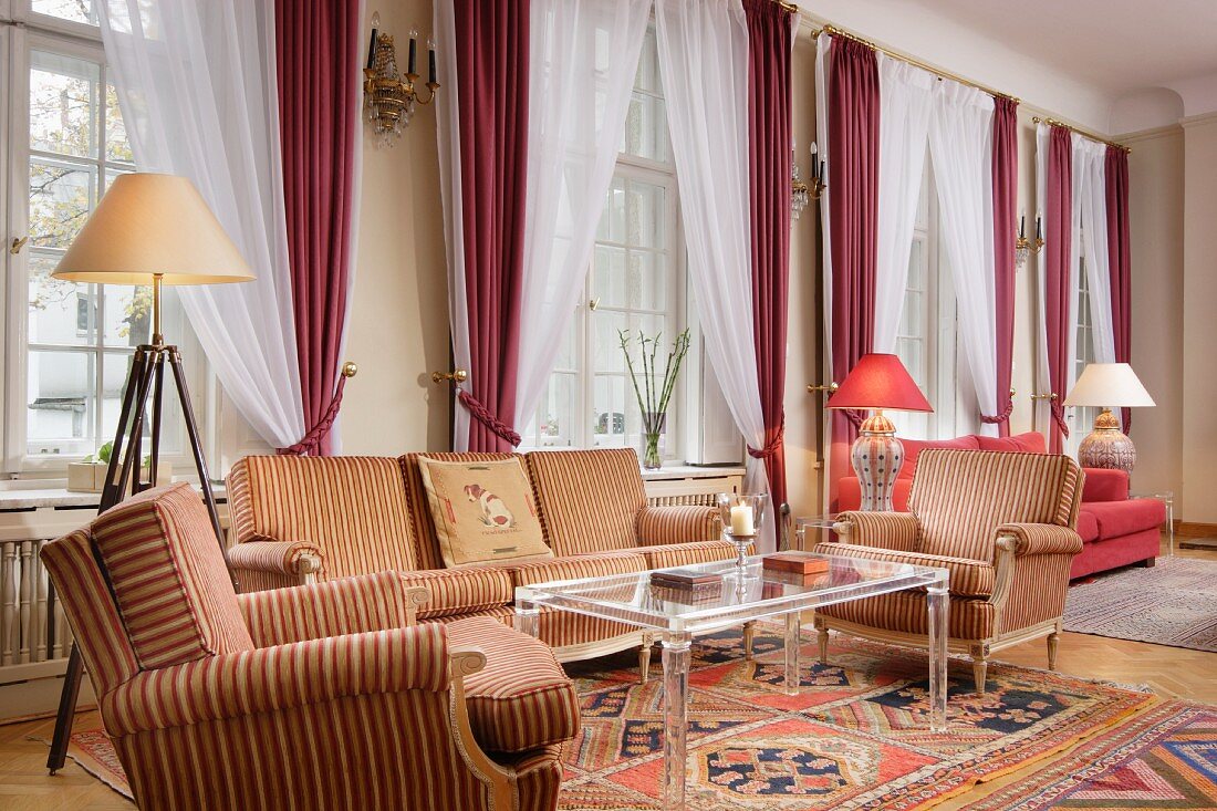 Grosszügiges Wohnzimmer mit gestreifter Polstergarnitur, Couchtisch aus Plexiglas & Gardinen mit rötlichen Vorhängen vor Sprossenfenstern