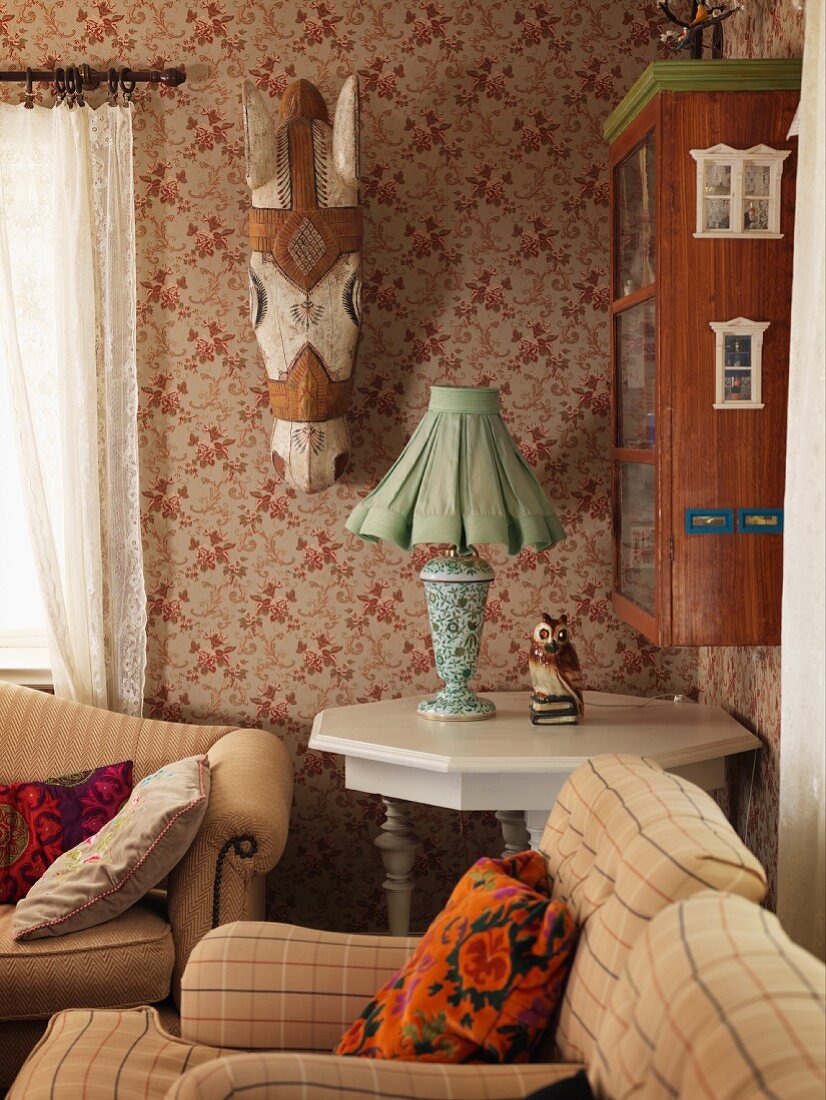 Polstersessel mit beigem Bezug, im Hintergrund Tischleuchte mit Stoffschirm auf Holztisch weiss lackiert, in Zimmerecke, an Wand bemalter Holz Pferdekopf