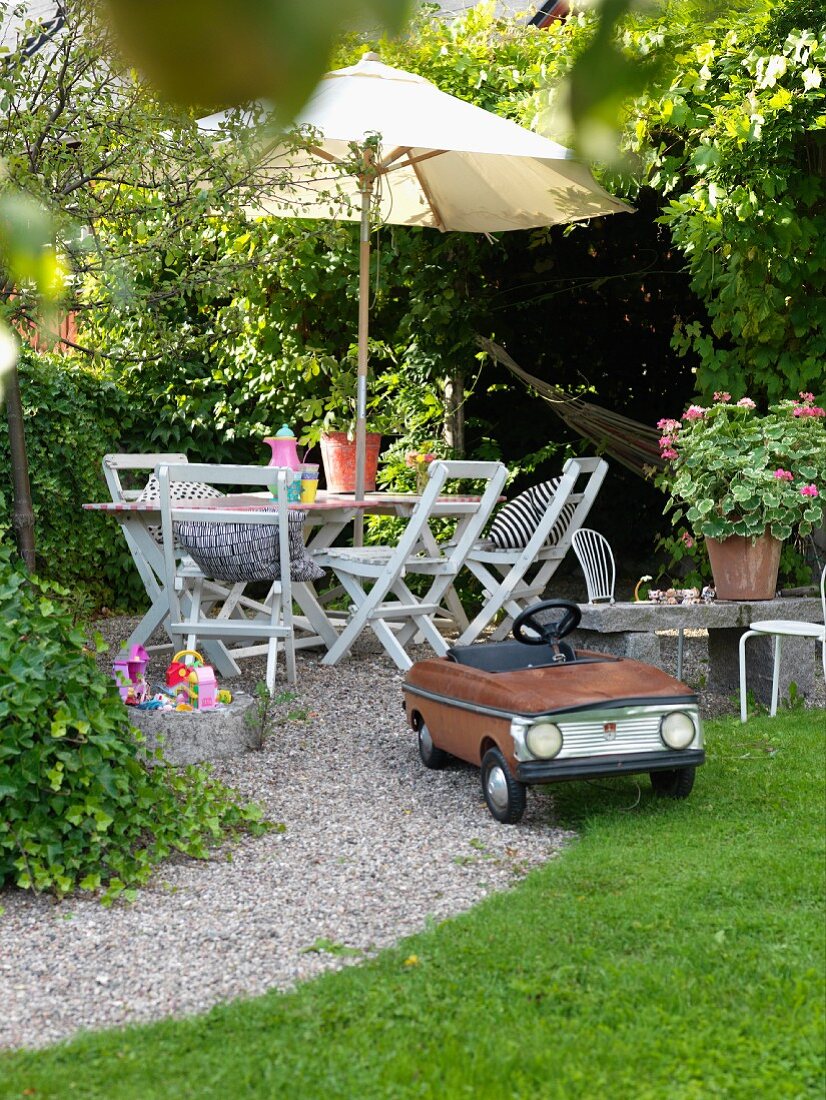 Retro-style pedal car on gravel path and idyllic seating area with table and chairs below parasol in garden