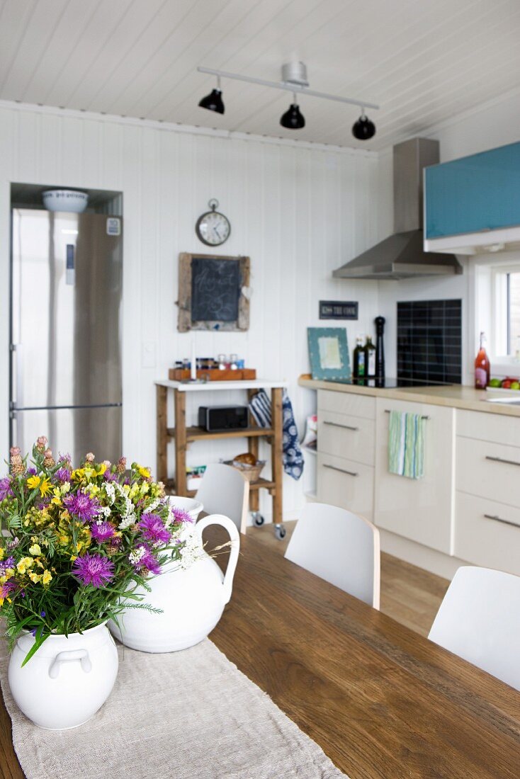 White vase of flowers on wooden table in front of kitchen counter against wood-clad white wall