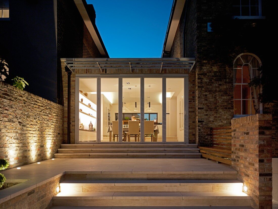 Steps leading to contemporary extension between two houses at night with view into illuminates dining room