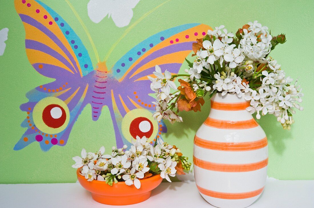 Weisser Flieder in Retro Keramikvase mit weissen und orangen Streifen sowie oranger Schale, dahinter Wand mit buntem Schmetterlingsmotiv